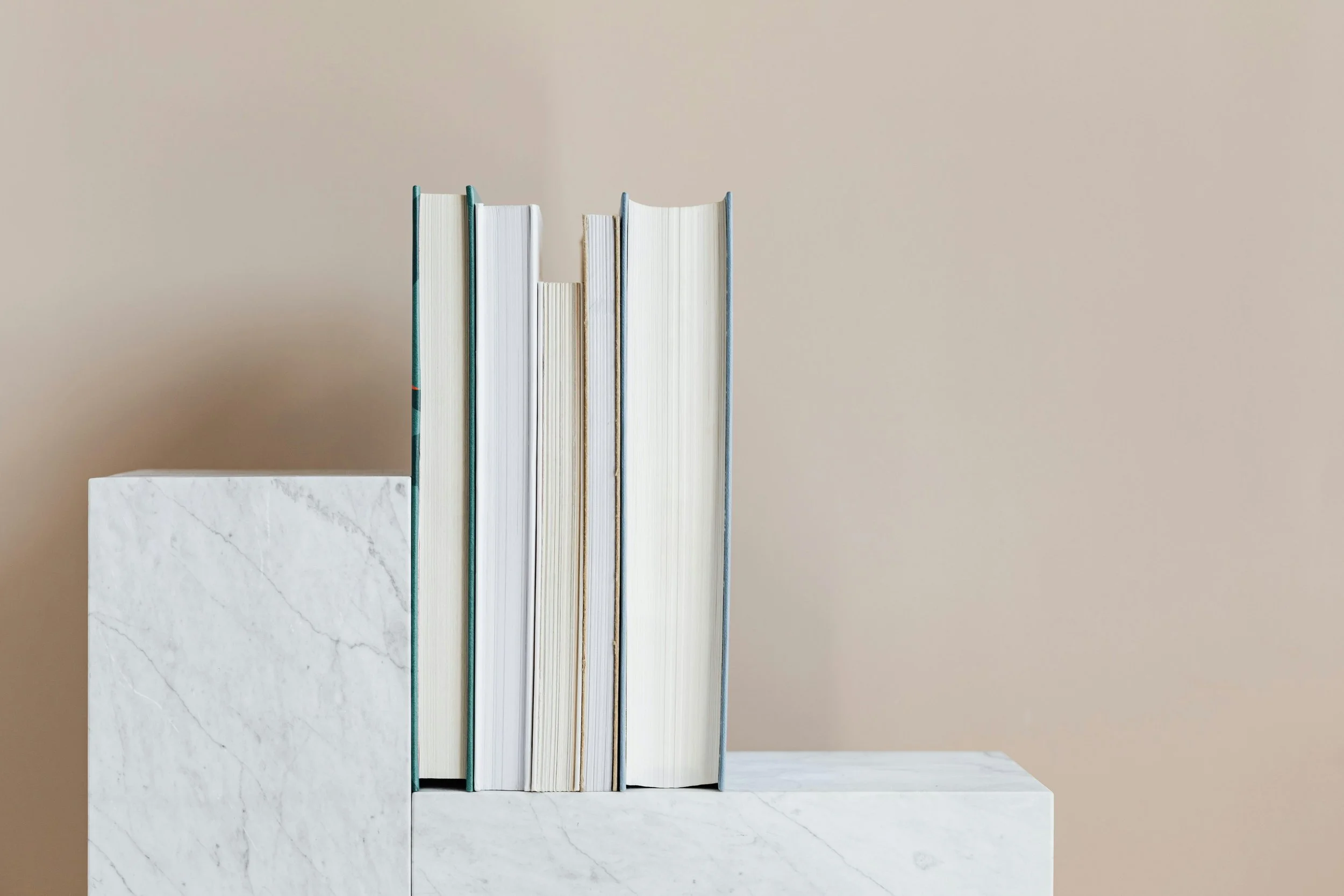 A row of six books of varying thickness standing upright on a white marble shelf against a plain beige wall.