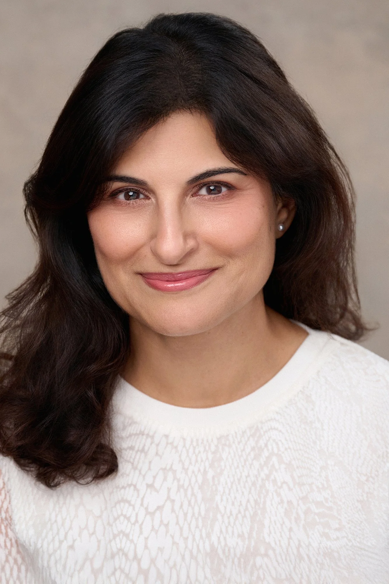 A woman with dark brown hair, wearing a white textured top and pearl earrings, smiling softly at the camera.