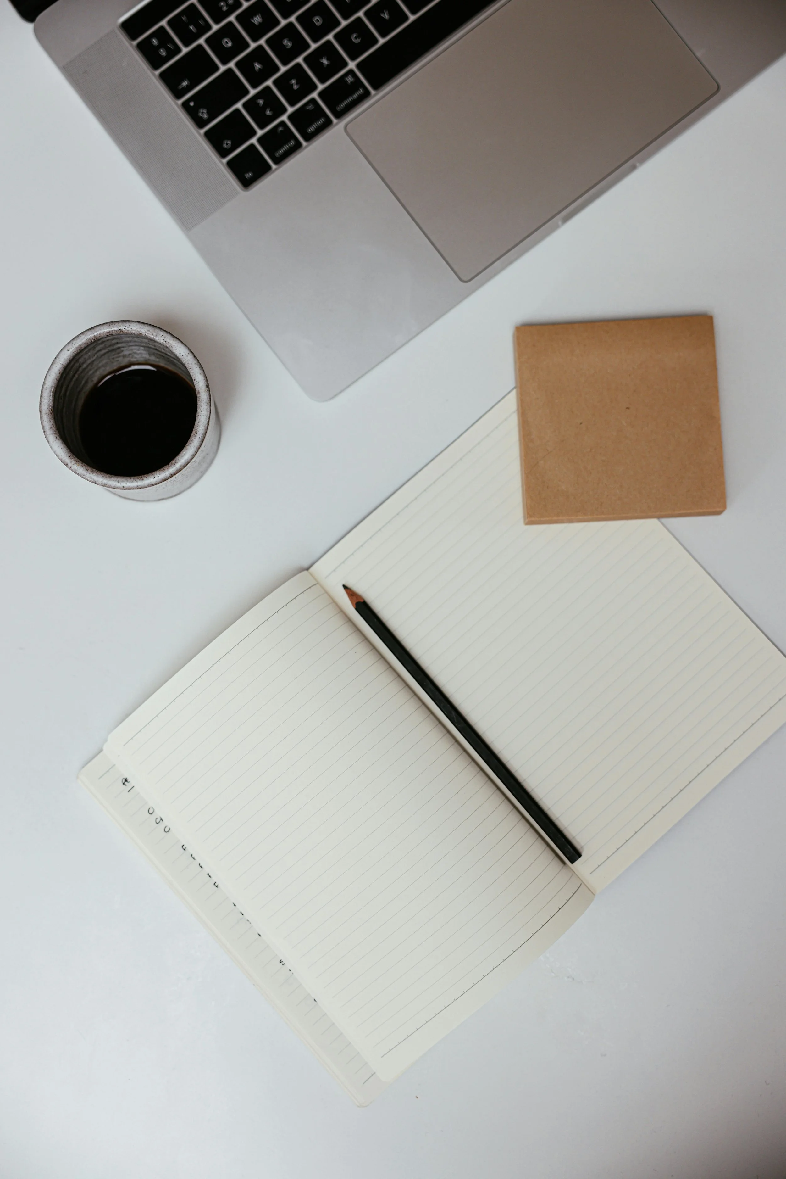 A workspace featuring a silver laptop, a cup of coffee, an open lined notebook with a black pencil, a small brown notebook, and a white surface.