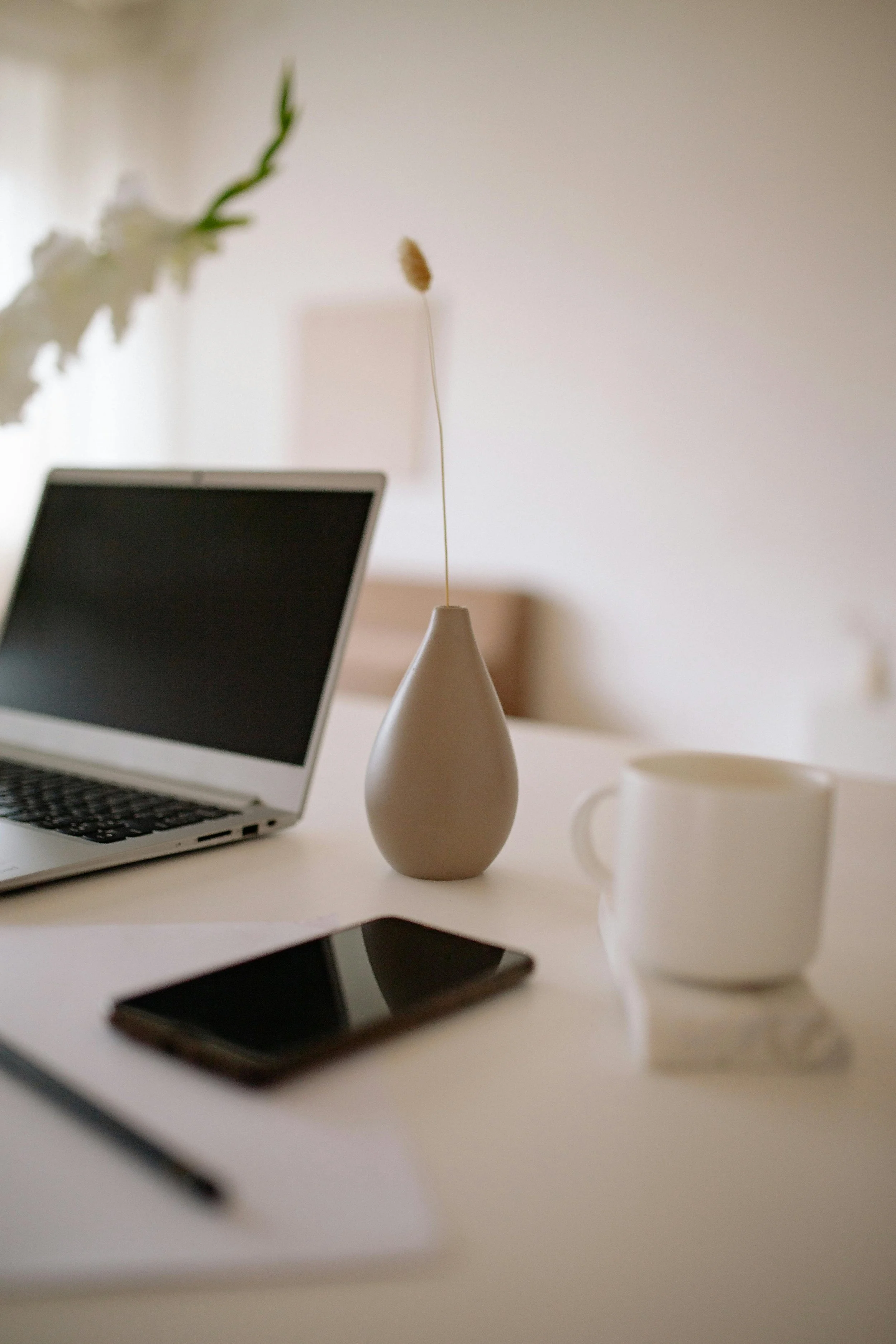 A minimalist workspace with a laptop, a smartphone, a white mug, a piece of paper, a pen, a beige vase with a dried flower, and a white flower in the background.