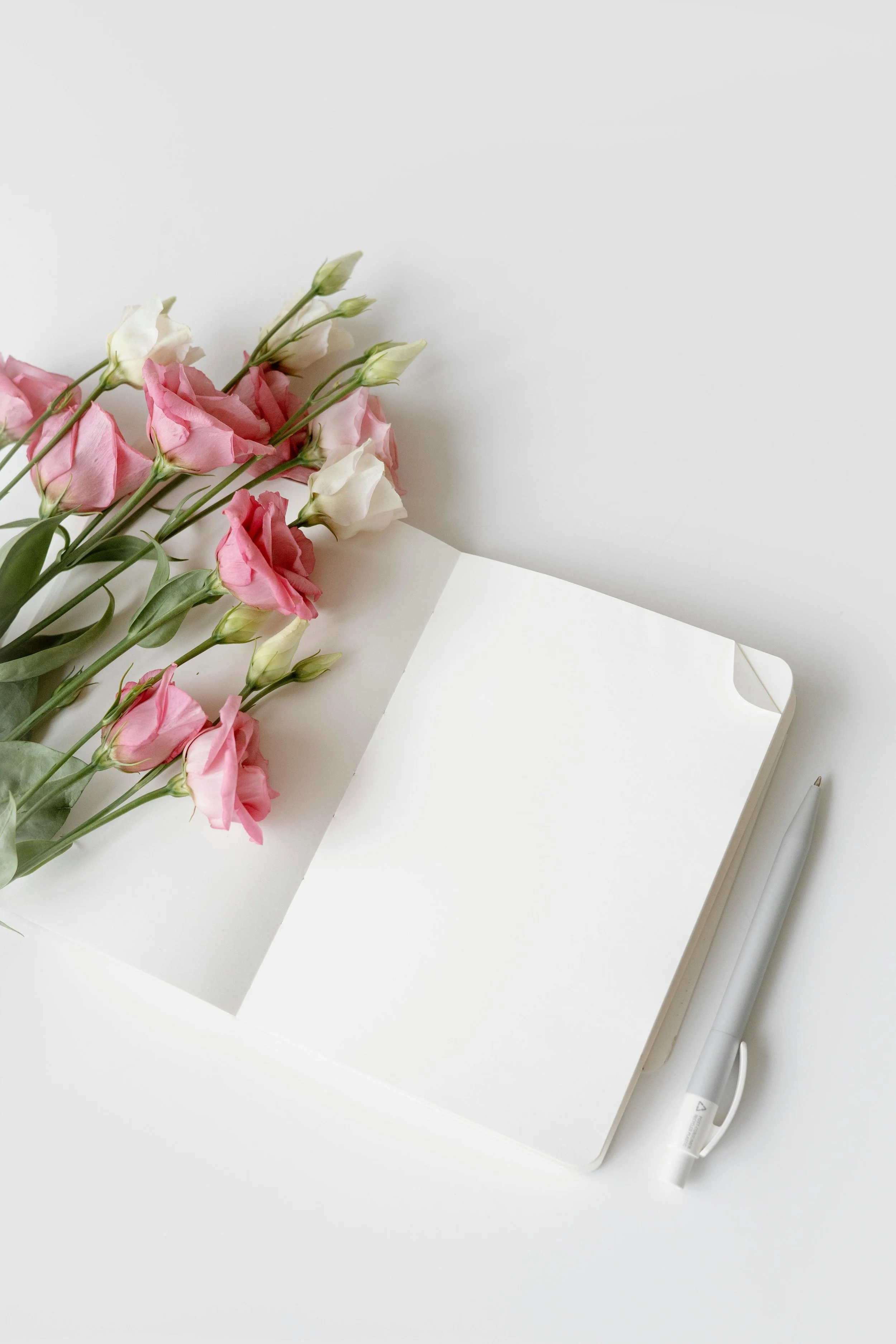 A white notebook, pink and white flowers, and a silver pen on a white surface.