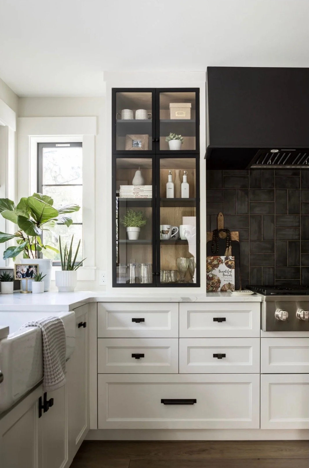 Kitchen with white cabinets, black accents, and glass-front storage cabinet with dishes and decorative items, next to a window with plants.