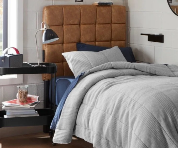 A cozy bedroom with a bed that has a brown padded headboard, gray and navy bedding, a bedside black table with a lamp, books, and a glass of water, and a white brick wall.