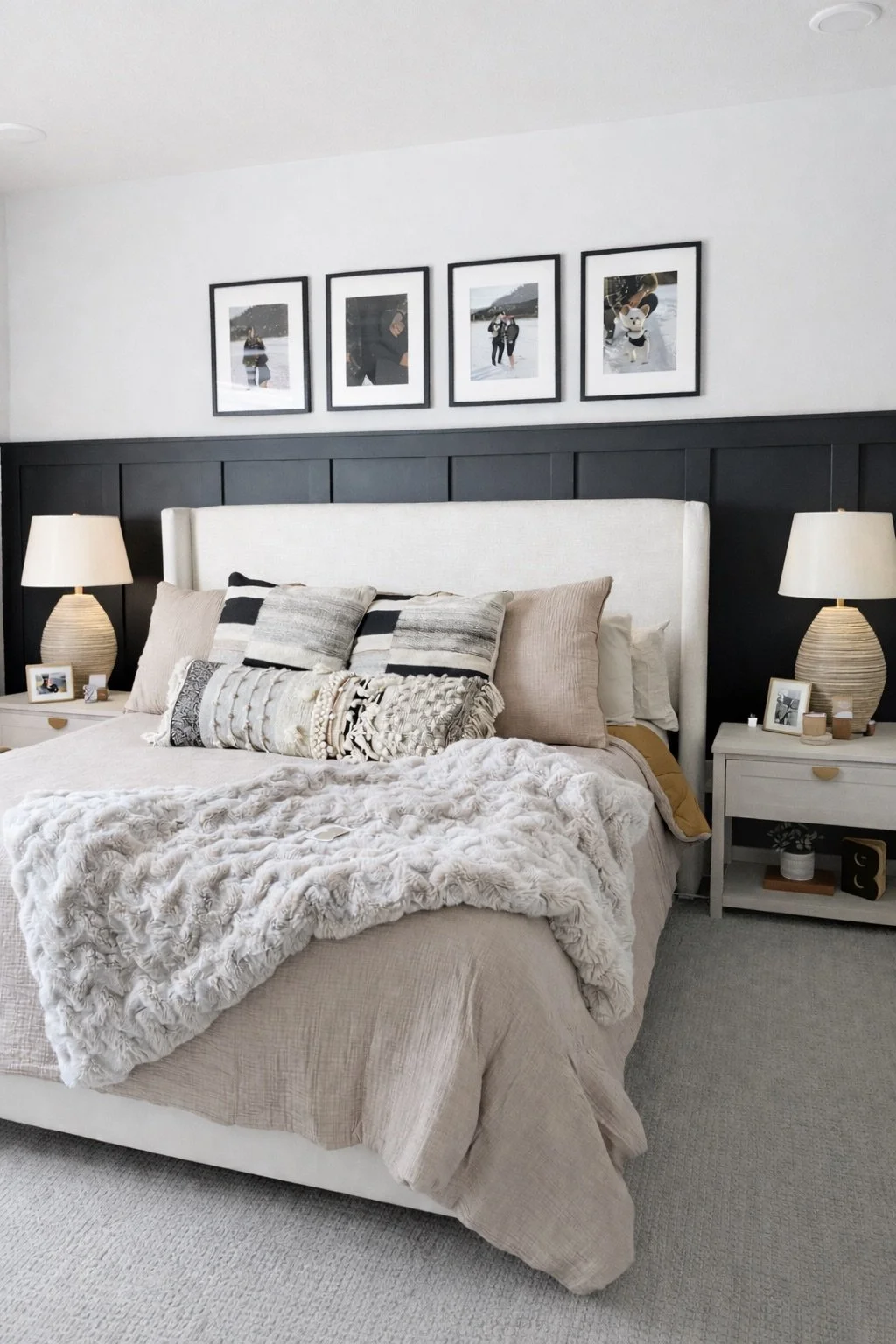 A neatly made bed with beige and cream bedding, decorative pillows, and a plush white blanket in a modern bedroom. Two matching nightstands with lamps and framed photos flank the bed. Above, four framed photos are hung on a white wall with a black wood-paneled stripe.