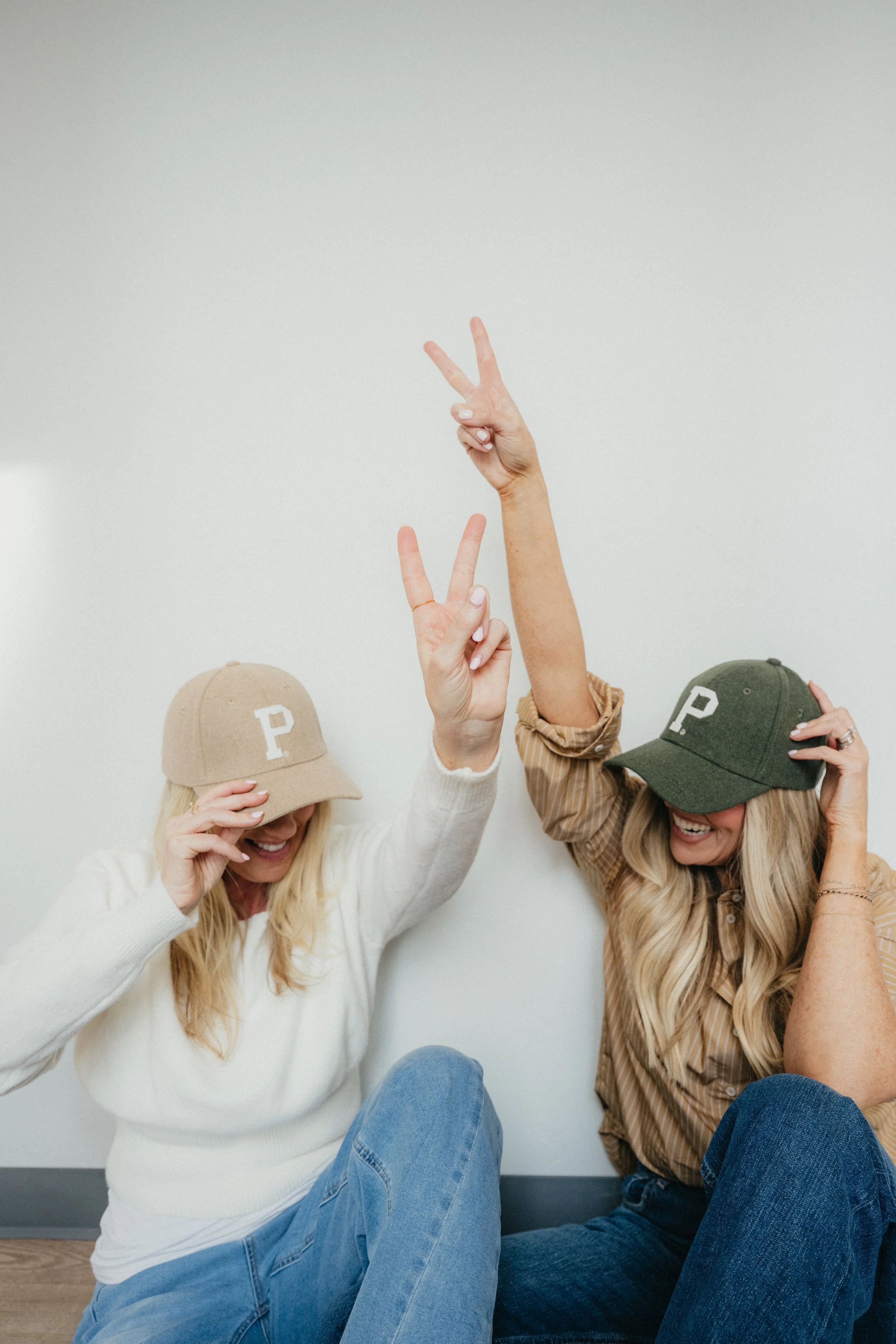 Two women sitting on the floor against a white wall, both wearing baseball caps with the letter 'P'; one woman is in a beige cap and white sweater, the other in an olive cap and a striped shirt. They are smiling and making peace signs with their hands, appearing happy and playful.