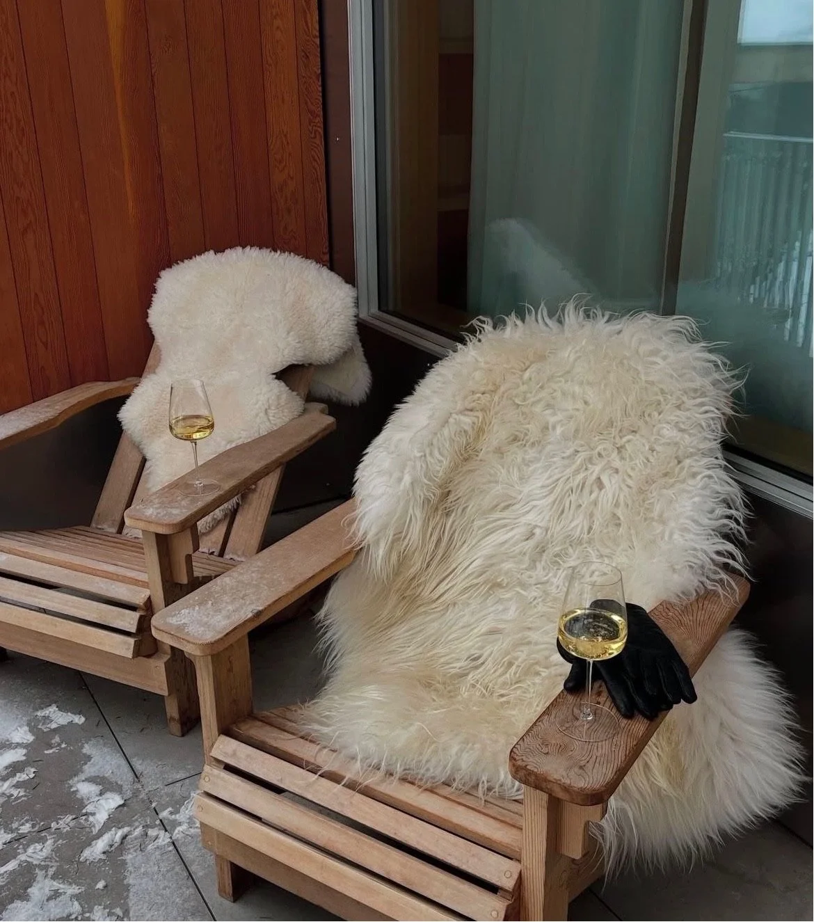 Two wooden Adirondack chairs with faux fur throws, each with a glass of white wine. One chair has a black glove laid on the armrest.