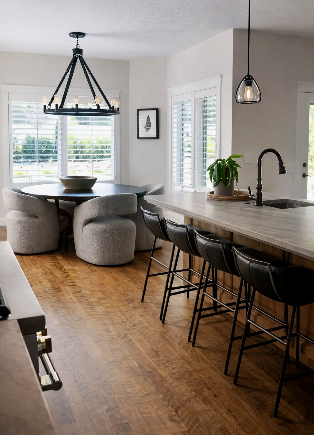 Modern kitchen and dining area with a round table and upholstered chairs, a kitchen island with bar stools, large windows with white blinds, wood flooring, and contemporary light fixtures.