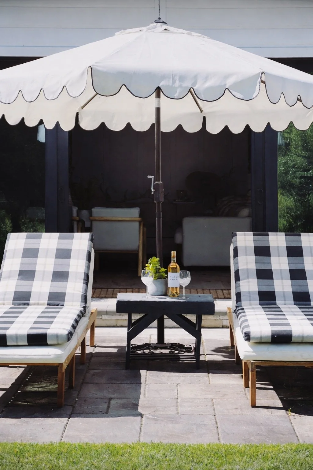 Outdoor patio with two checkered lounge chairs, a small black table with a plant and a wine bottle, beneath a large white patio umbrella, with a sofa and windowed door in the background.