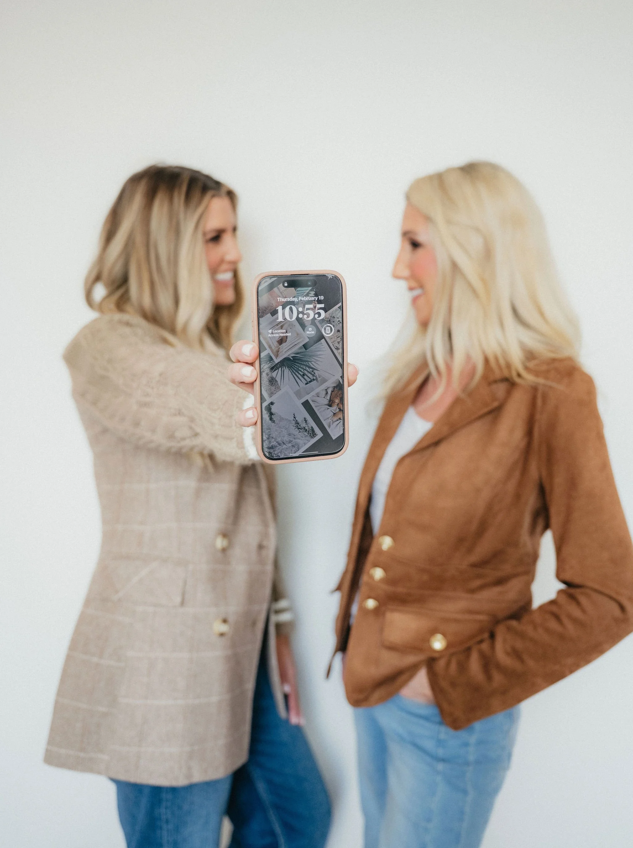Two women with blonde hair, one holding a smartphone, are standing against a plain white wall, smiling and facing each other.