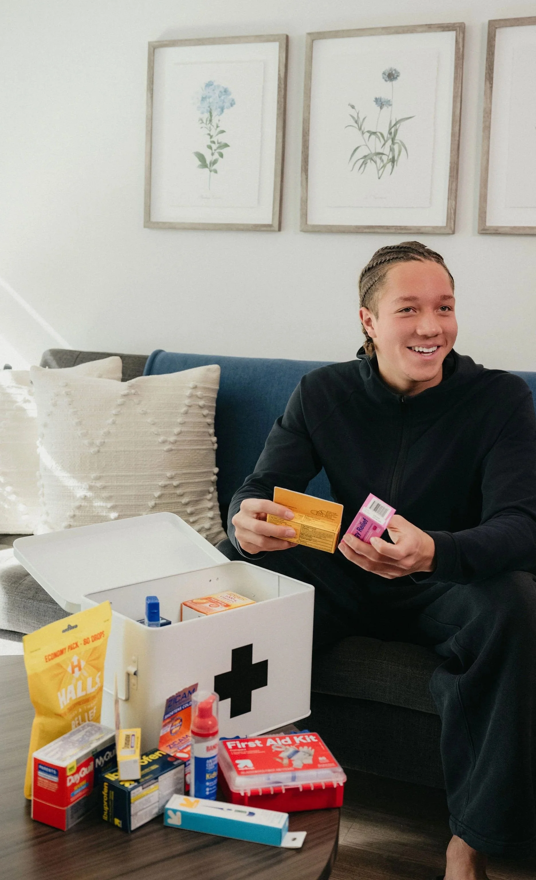 Young man with braided hair sitting on a couch, smiling, holding medication boxes, surrounded by a first aid kit and various medical supplies on a table.