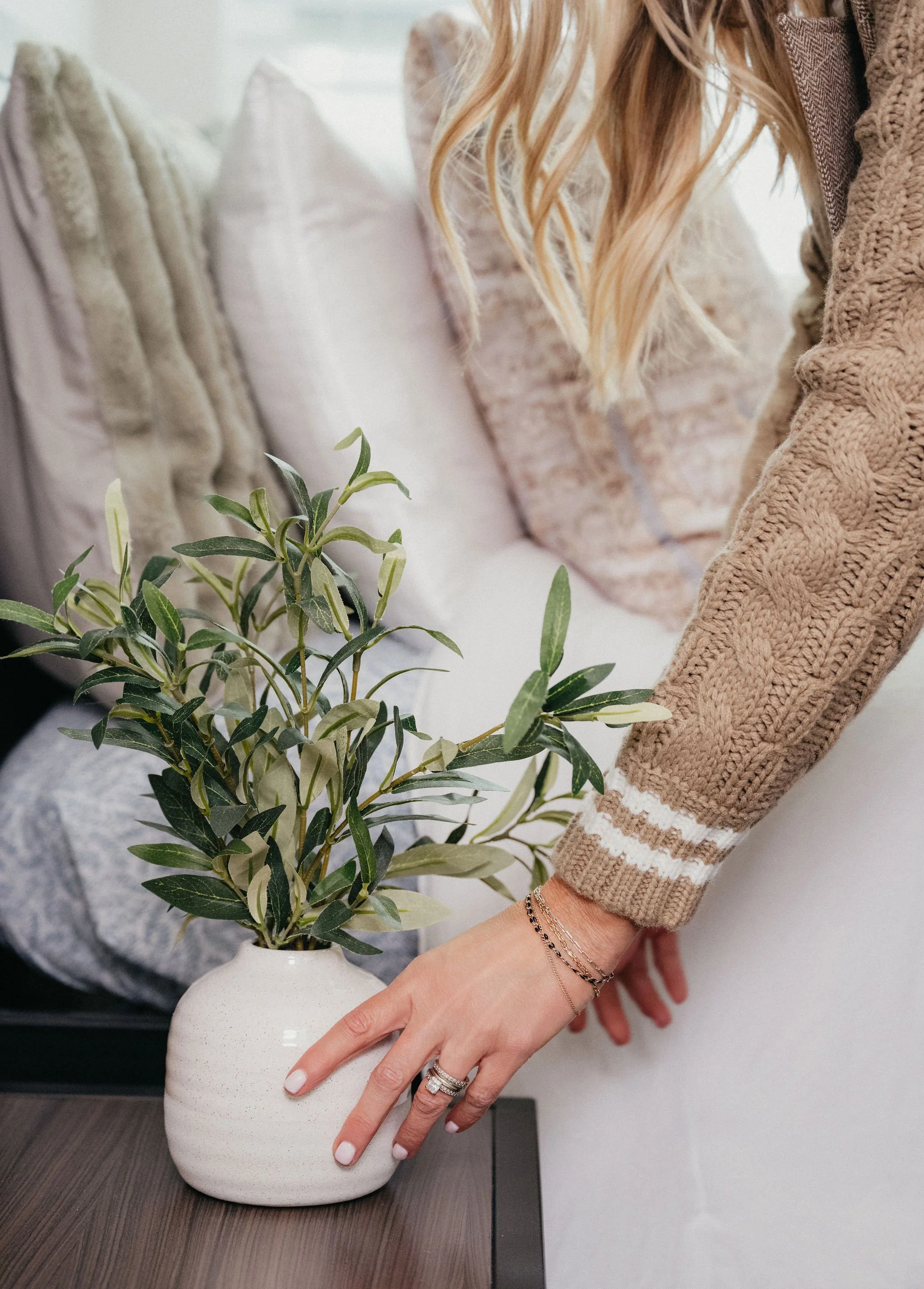 A woman with long blonde hair wearing a tan cable-knit sweater and jewelry, placing her hand on a white ceramic vase with green leafy plant on a wooden table.