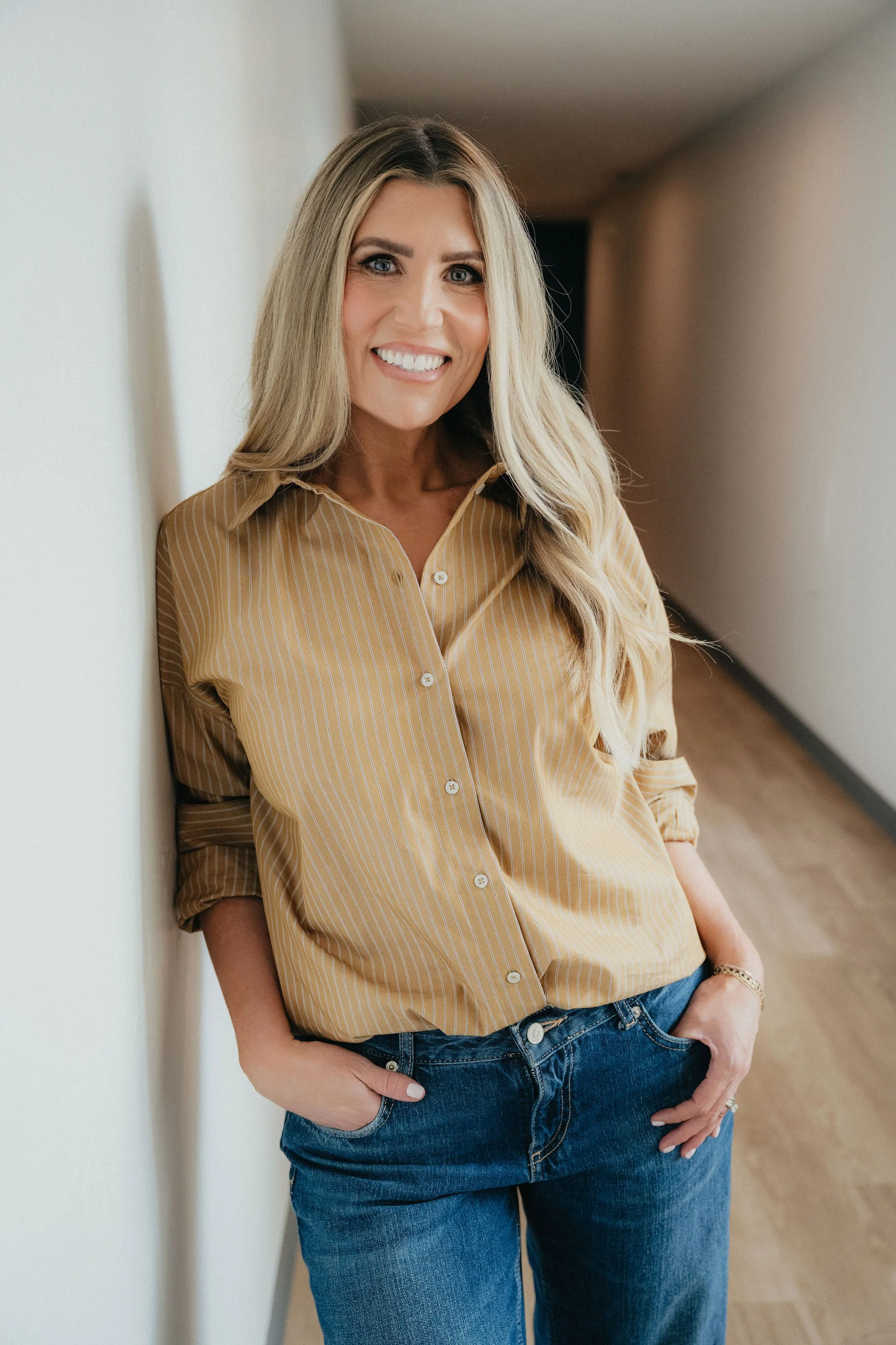 A woman with long blonde hair, wearing a beige striped button-up shirt and blue jeans, smiling and leaning against a wall in a hallway.