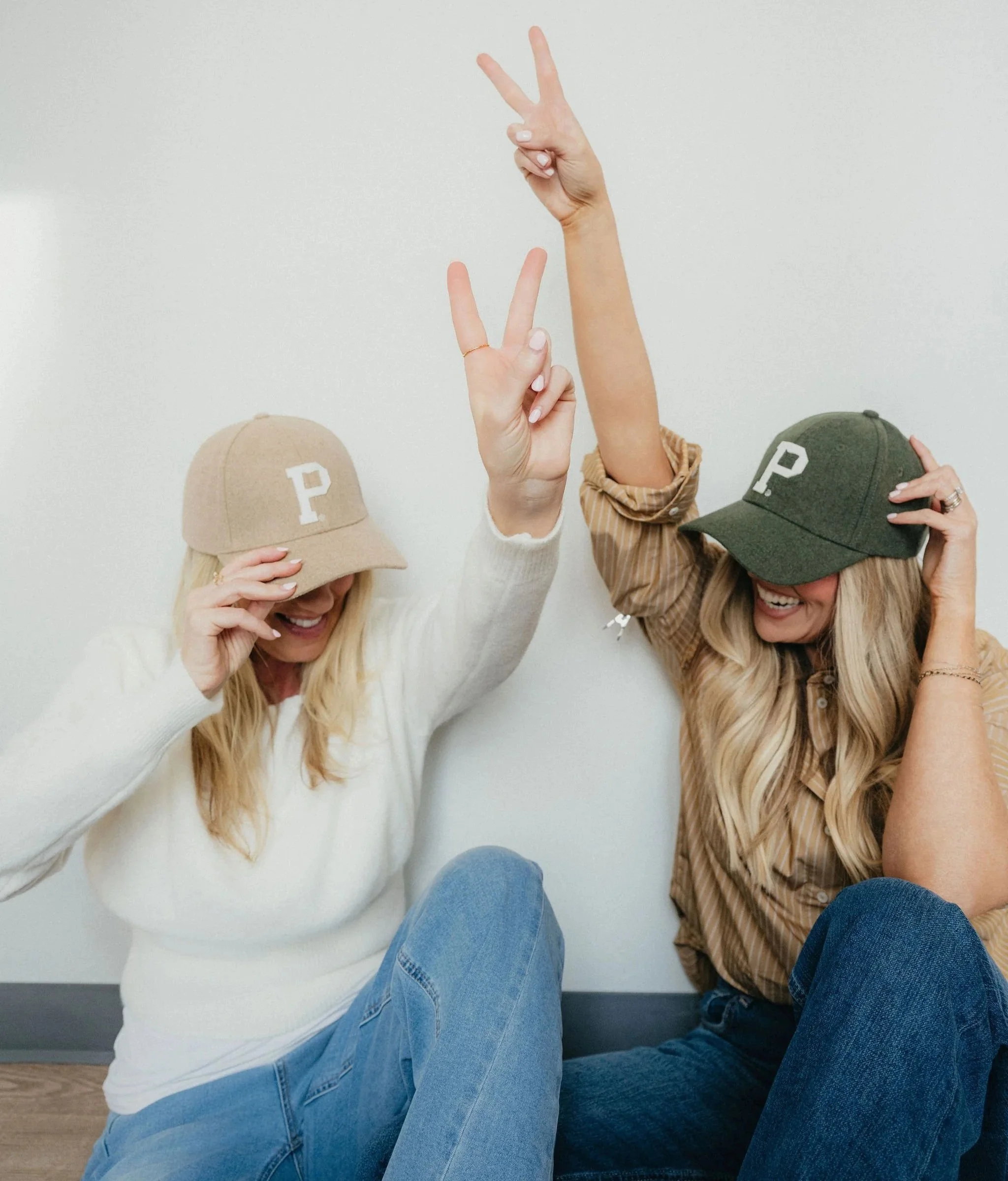 Two women sitting on the floor, wearing caps with a 'P' on them, smiling, and making peace signs with their hands.