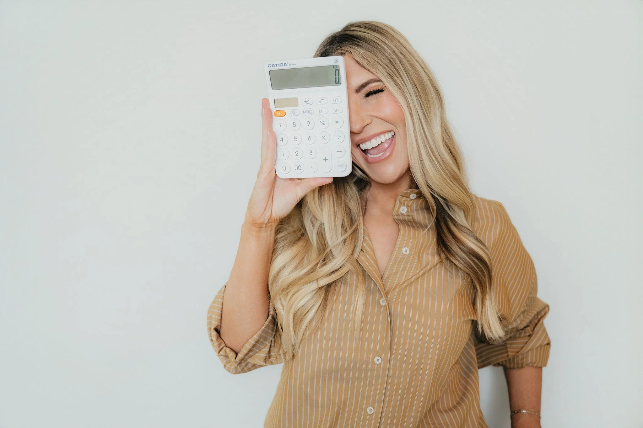 A woman with long blonde hair smiling and holding a calculator up to her face with her right hand, covering part of her face, standing against a plain light background.