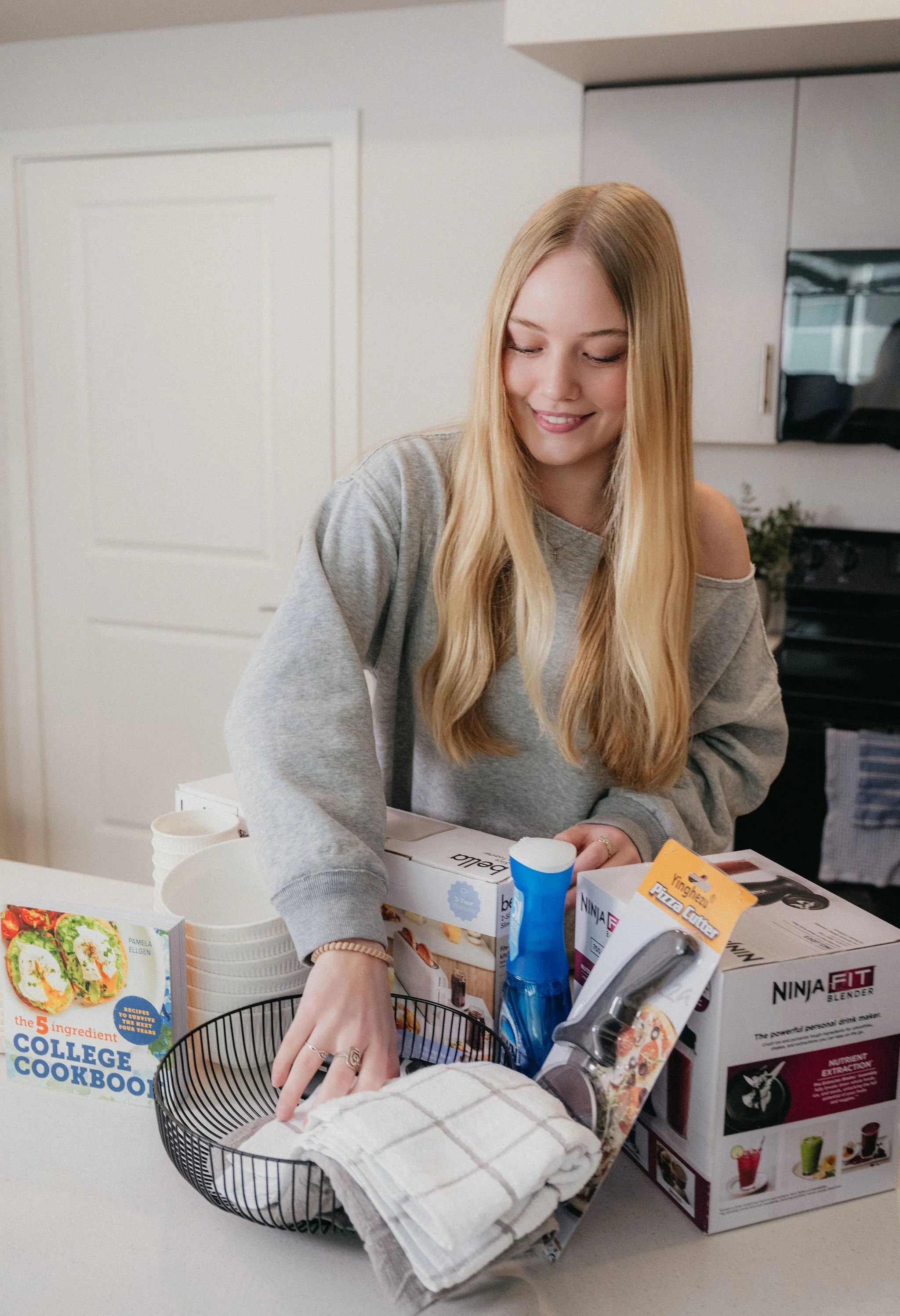 A young woman with long blonde hair wearing a gray sweatshirt standing in a kitchen. She is smiling and reaching into a black wire basket containing a white dish towel. On the counter are various kitchen items including a Ninja blender, a box, a cleaning brush, and a cookbook titled 'The 5 Ingredient College Cookbook.'