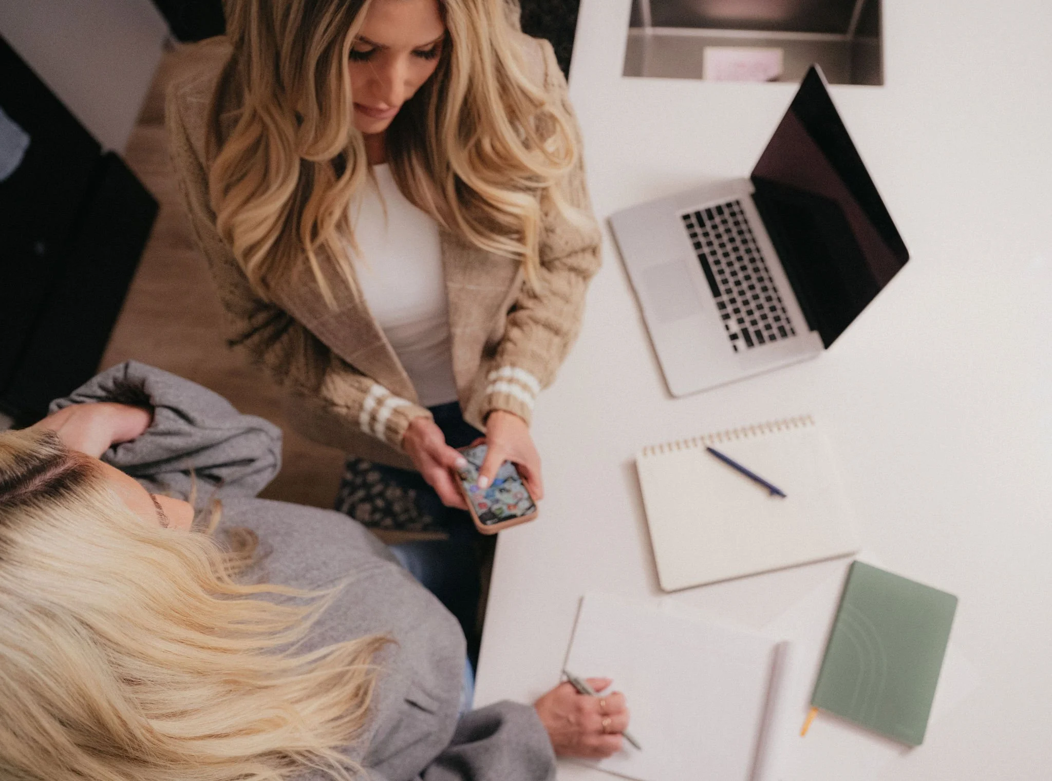 Two women working at a white table with laptops, notebooks, and a pen, one is holding a smartphone, in an office setting.