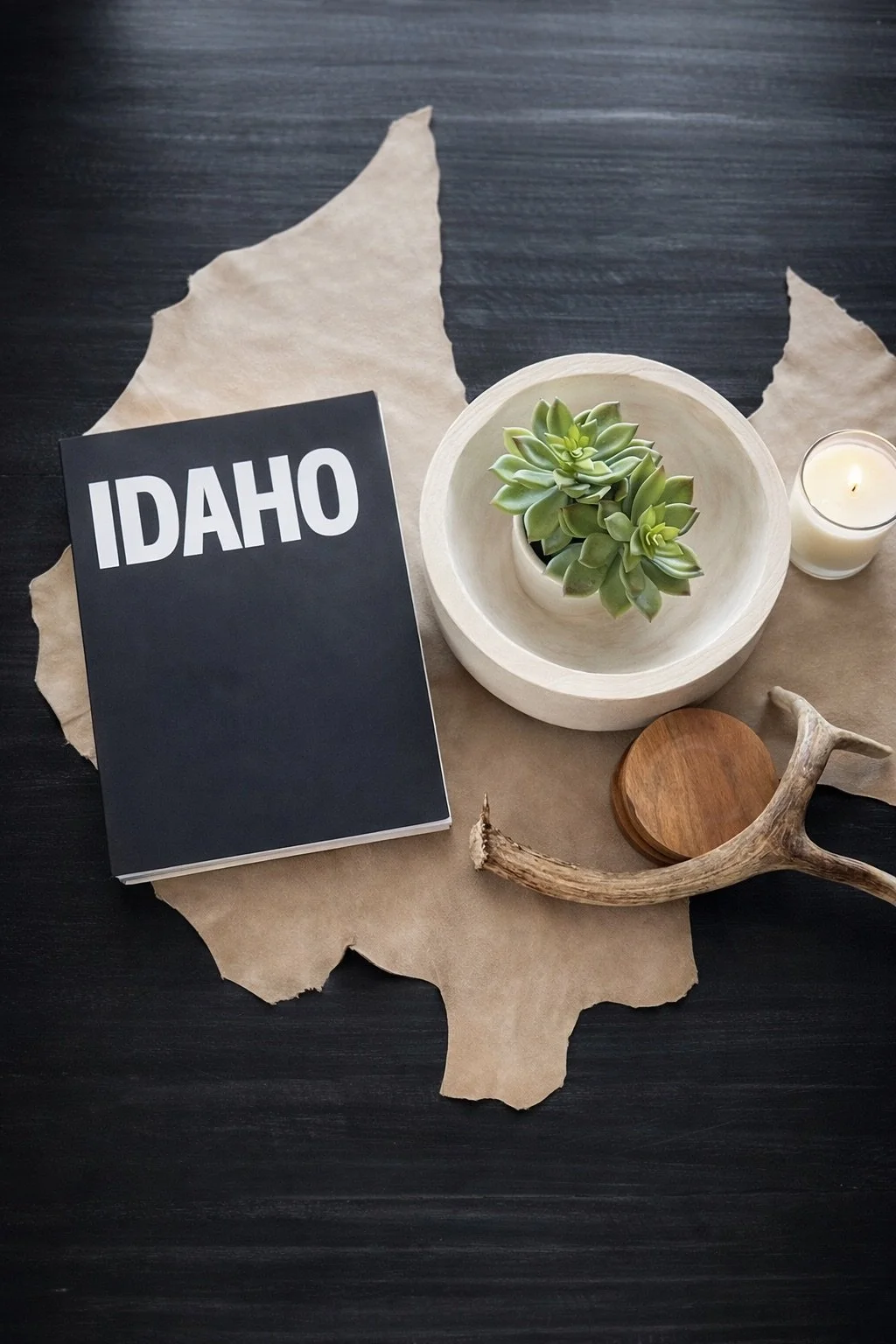 A flat lay of decorative items on a dark wooden surface, including a black and white Idaho notebook, a white bowl with green succulents, a white candle in a glass holder, a piece of torn brown paper, a wooden coaster, and a natural antler piece.