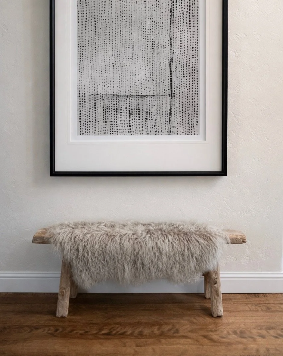A wooden bench with a sheepskin rug on top, positioned below a framed black-and-white abstract artwork on a white wall.