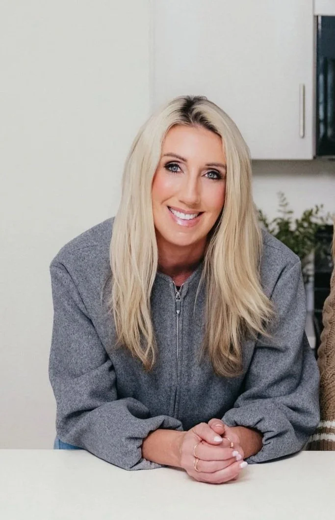 A woman with long blonde hair, wearing a gray zip-up sweater, smiling and sitting at a table in a bright room.