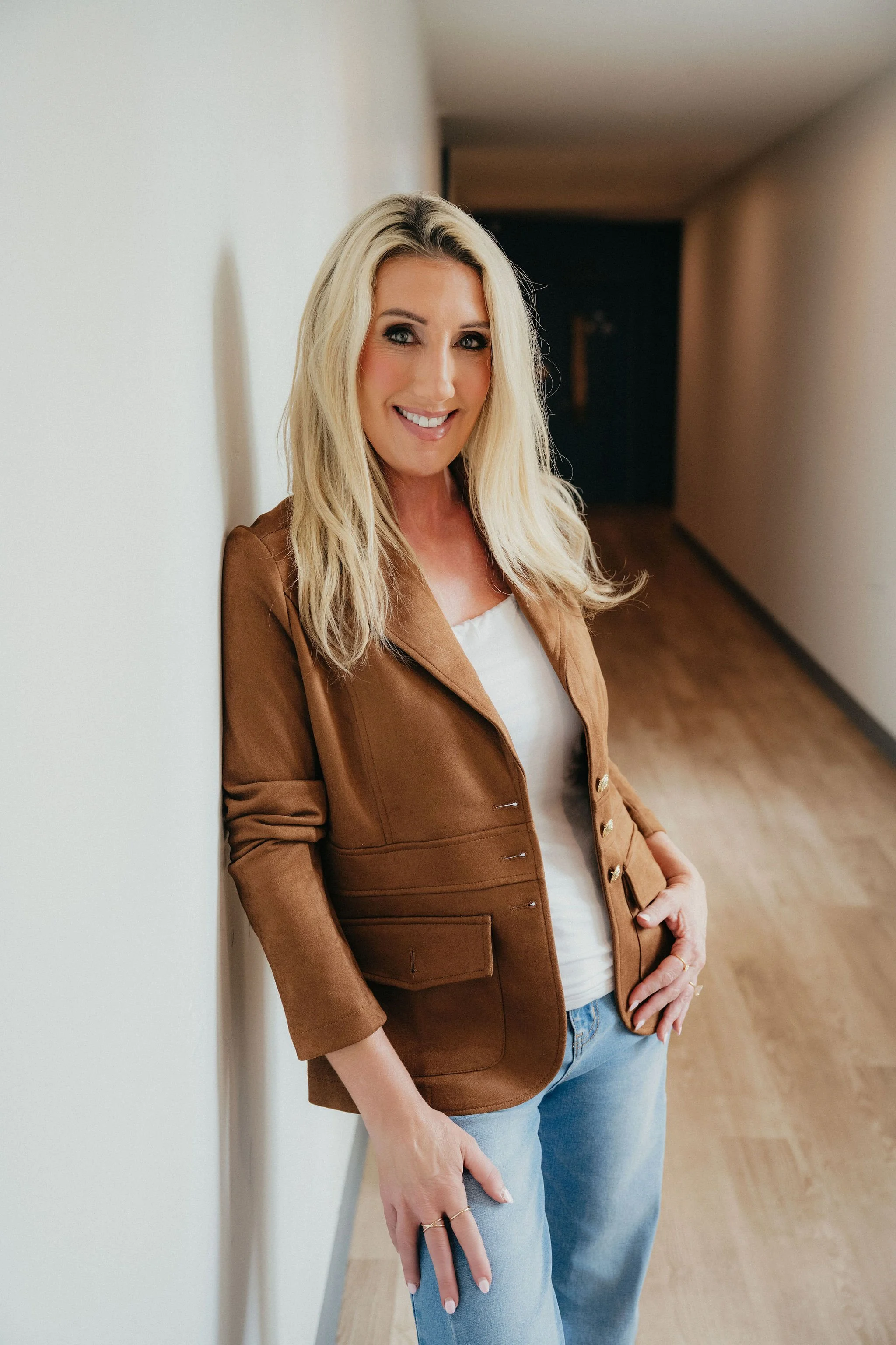 A smiling blonde woman leaning against a white wall in a hallway, wearing a brown jacket, white top, and light blue jeans.