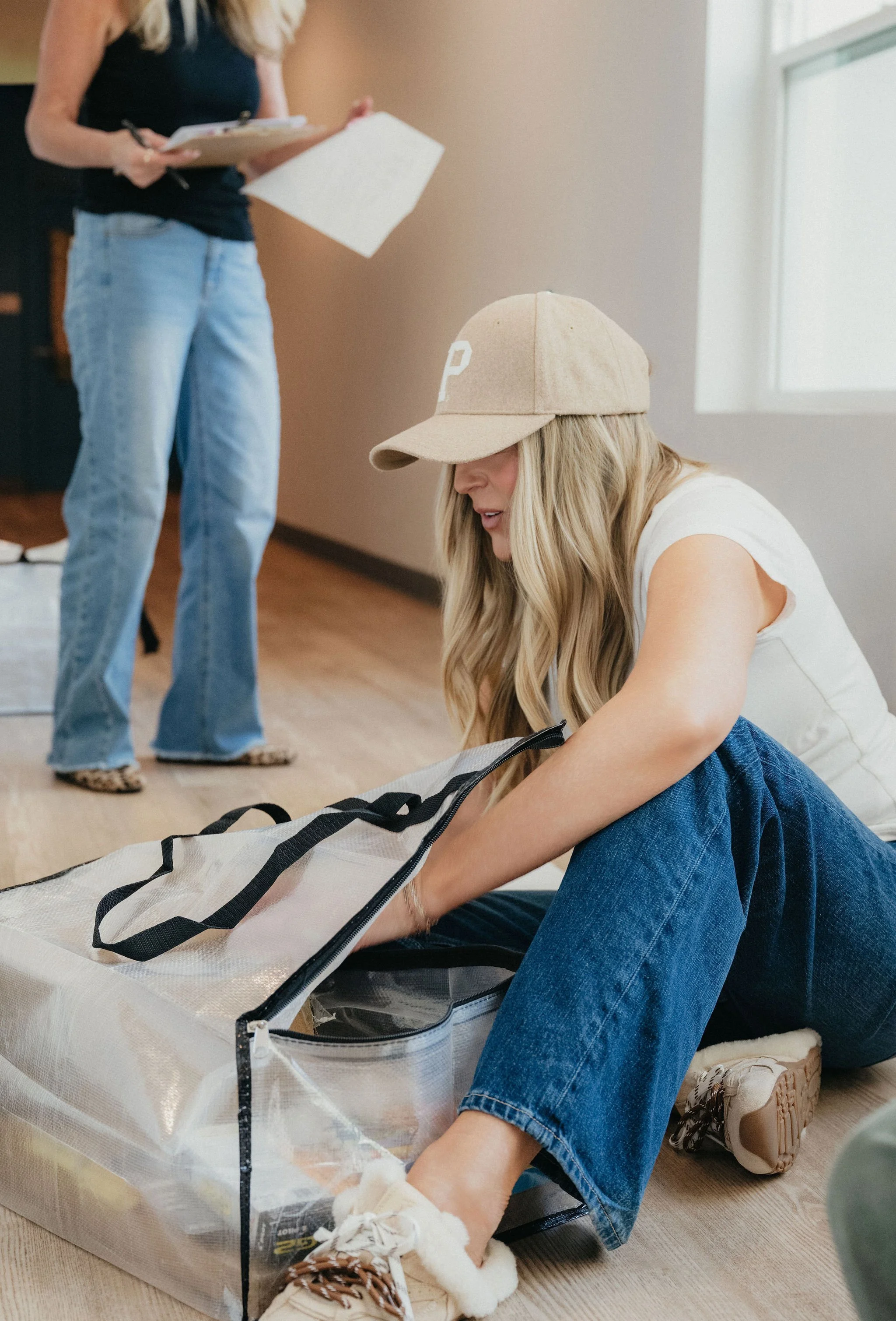 A woman with long blonde hair, wearing a tan cap, white t-shirt, and blue jeans, is sitting on the floor packing a transparent bag. In the background, another woman is standing, holding a clipboard and pen, wearing a black tank top and light blue jeans.