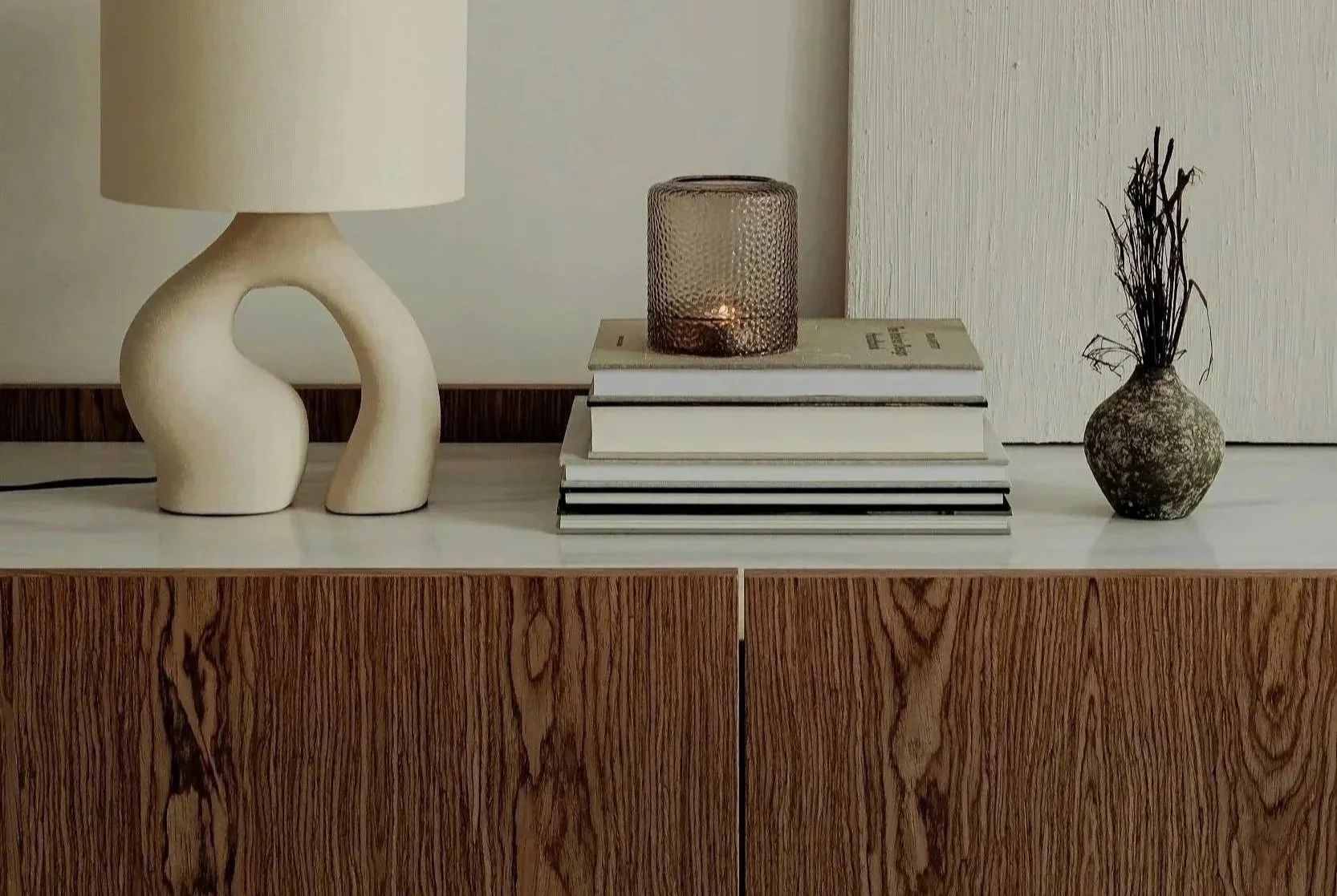 Decorative table with a beige lamp, a stack of books with a candleholder on top, and a small vase with twigs.