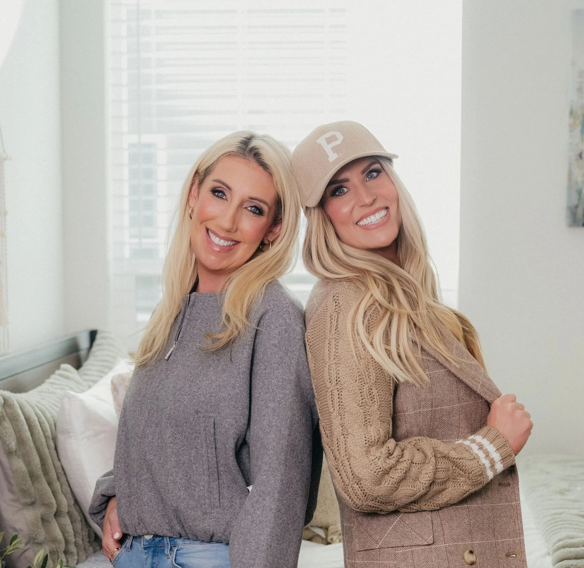 Two women with long blonde hair smiling and posing back-to-back indoors near a bed with pillows, in a bright room with large windows and white shutters.