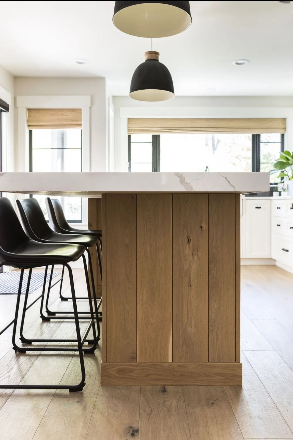 Modern kitchen with a wooden island, white marble countertop, black bar stools, large windows with blinds, and black pendant lights.