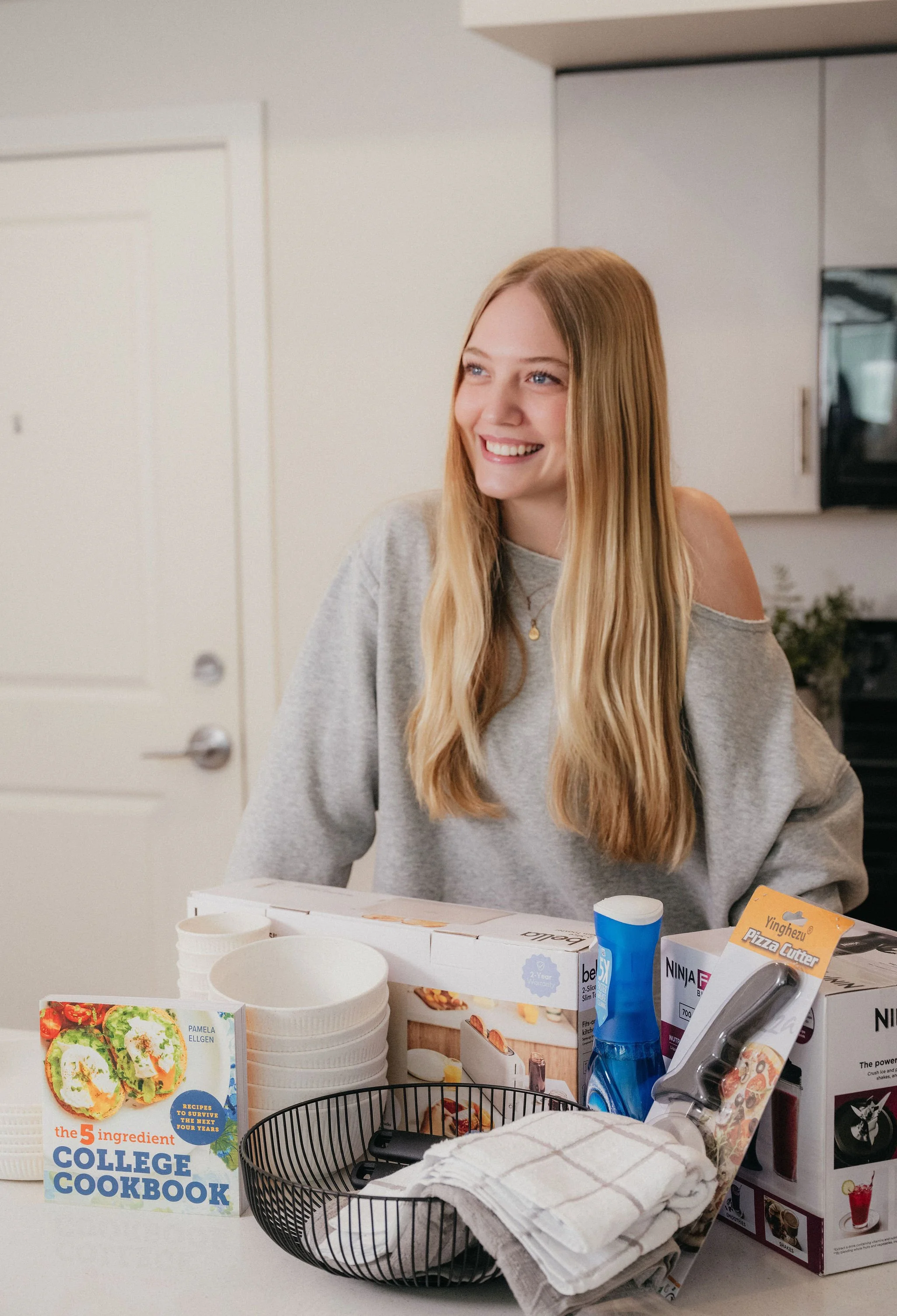 A young woman with long blonde hair smiling in a kitchen, surrounded by kitchen items such as bowls, a pizza cutter, and a cookbook titled '5 Ingredient College Cookbook'.