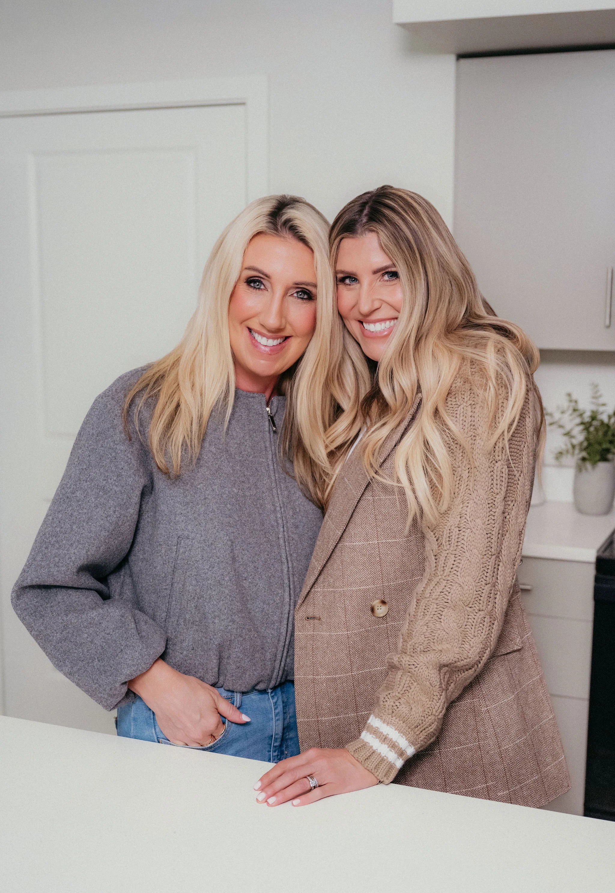 Two women smiling and embracing in a modern kitchen, standing behind a white countertop.