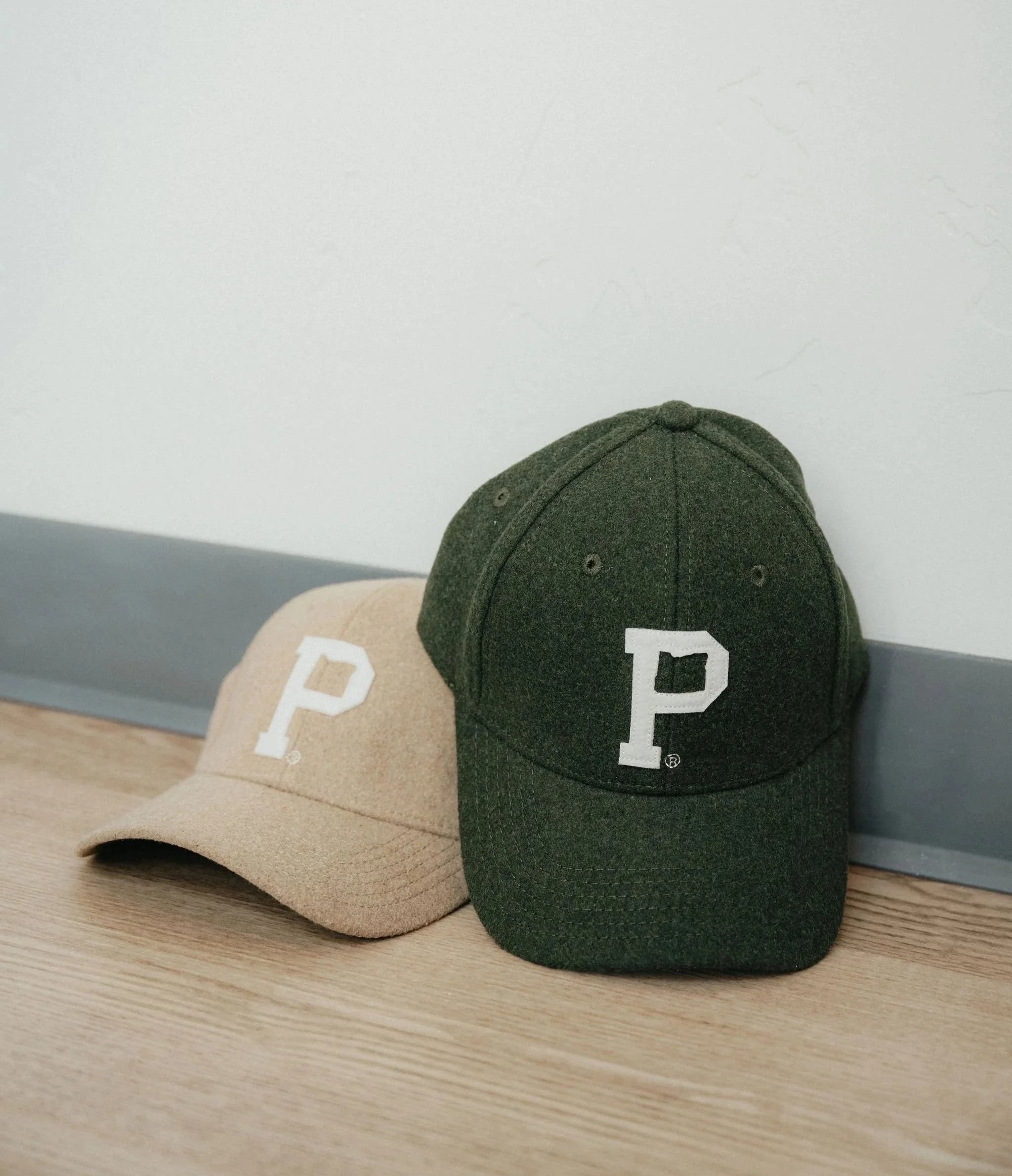 Two baseball caps, one beige and one dark green, both with a white letter 'P' embroidered on the front, placed on a light wooden surface against a white wall.