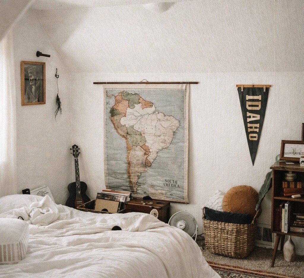 A bedroom with a bed in the foreground, a black acoustic guitar leaning against the wall, a wall map of South America, a Boise State University pennant, and a wicker chair with a cushion. There are other items like books, a fan, and framed photos.