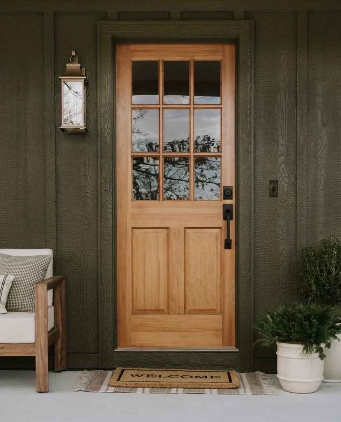 Front door made of light wood with glass window panes, framed by dark green textured wall featuring a lantern-style light fixture on the left and potted greenery on the right, with a welcome doormat on the floor.