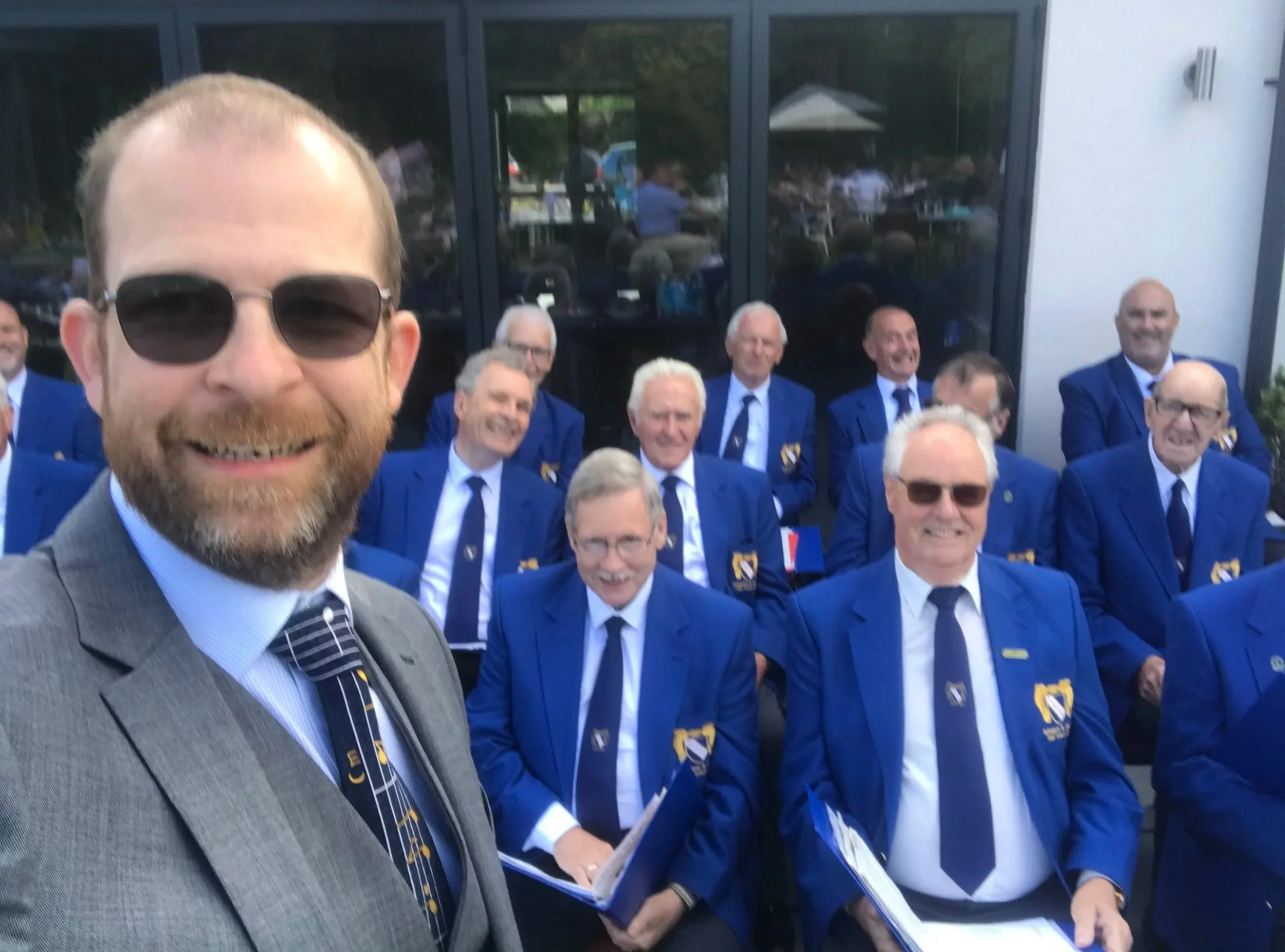 Man in suit, tie, and sunglasses taking selfie with group of men in blue blazers with yellow accents, outside building with glass windows.