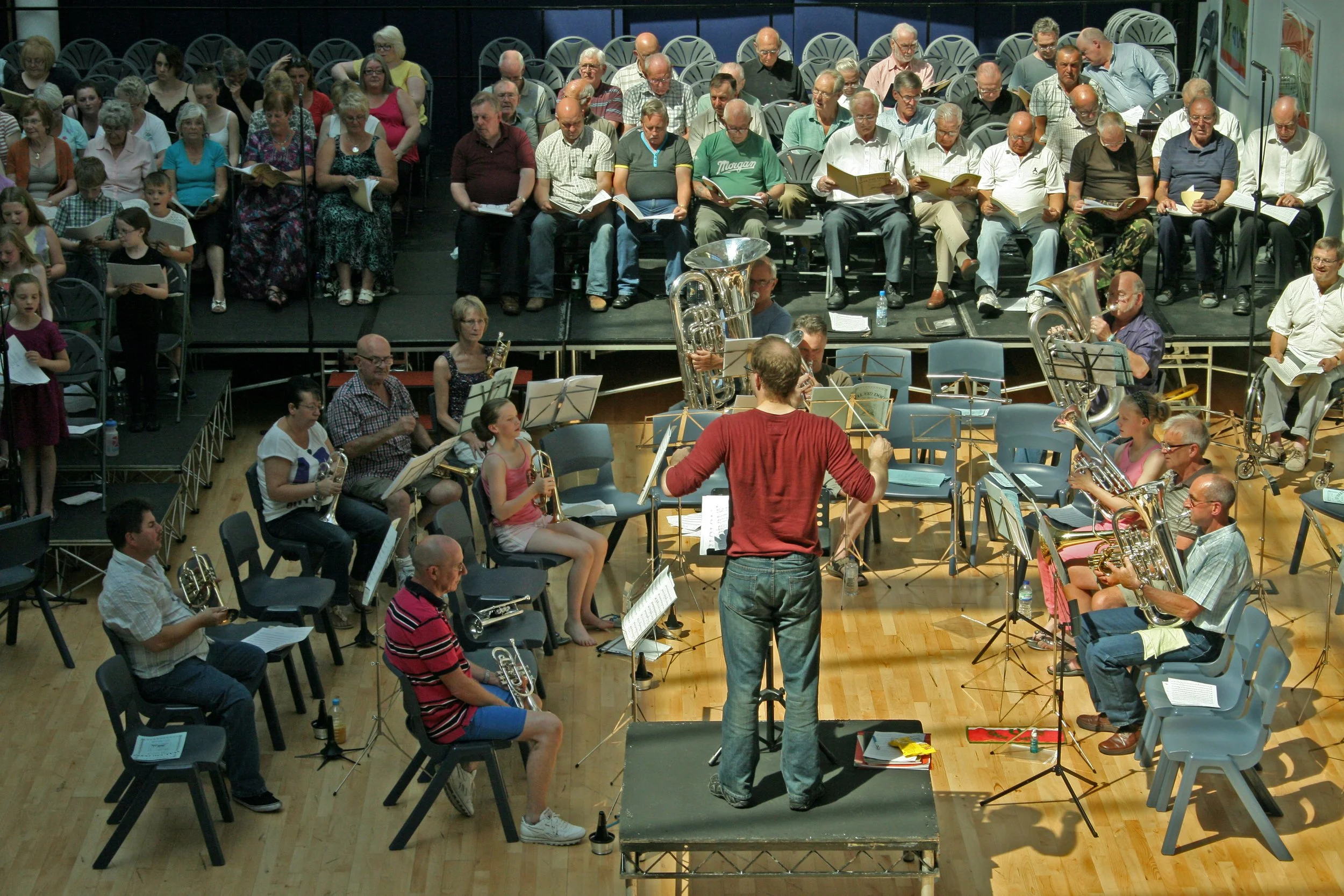 An indoor concert hall filled with choir singers and a small orchestra, with a conductor leading them.