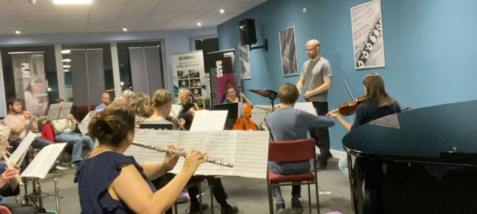 Orchestra performing in a room with a conductor leading the musicians, who are playing various instruments including flutes, violins, and a trumpet, with blue walls and framed artwork.
