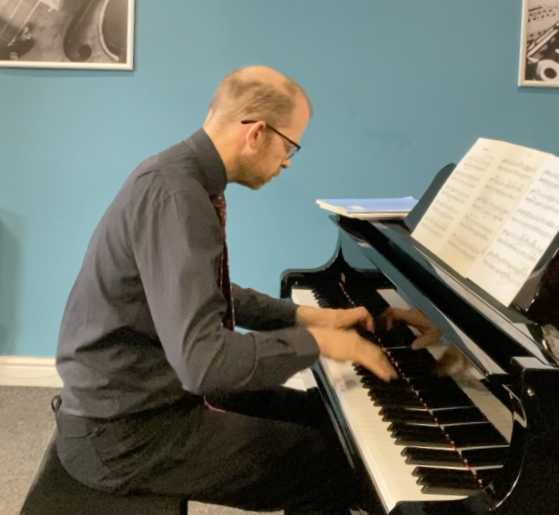 A man with glasses, dressed in a dark shirt and tie, playing a grand piano in a room with blue walls and framed pictures.