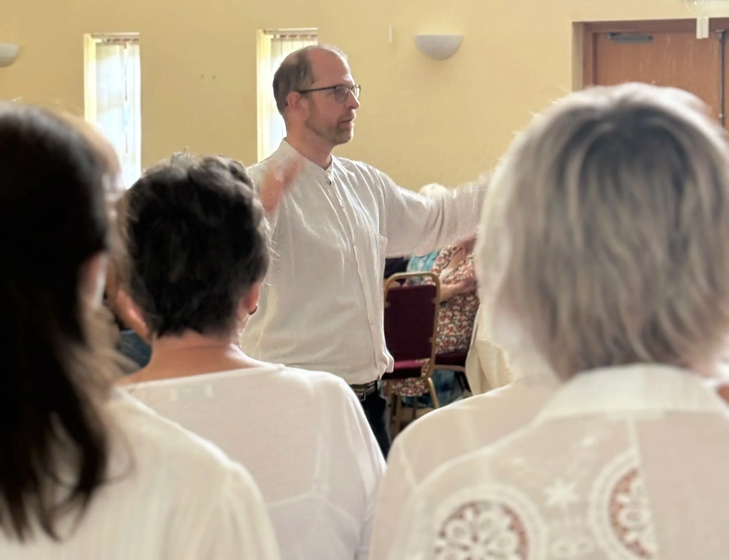 A man with glasses and a white shirt standing in front of a group, gesturing with one arm extended, in a setting with yellow walls and windows.