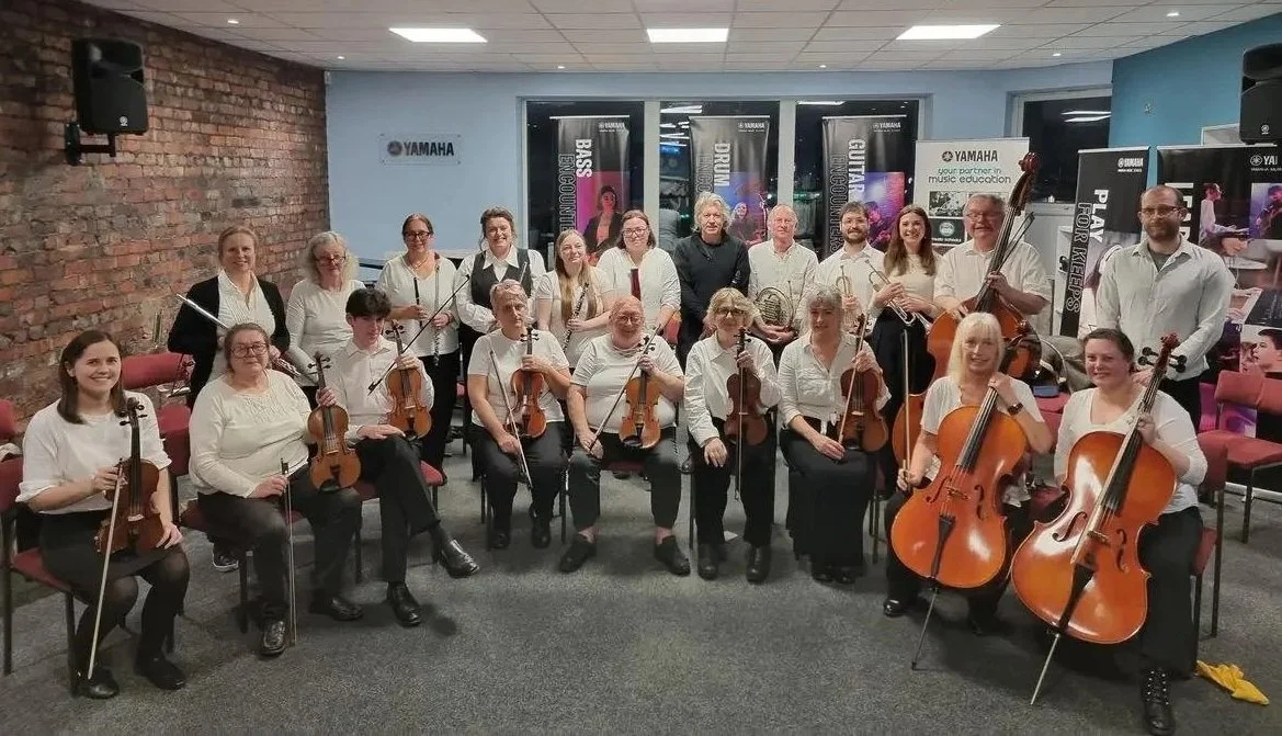 Group photo of a large string orchestra with members holding violins, cellos, and violas, in a room with brick and painted walls, some standing and some seated, all dressed in formal attire.
