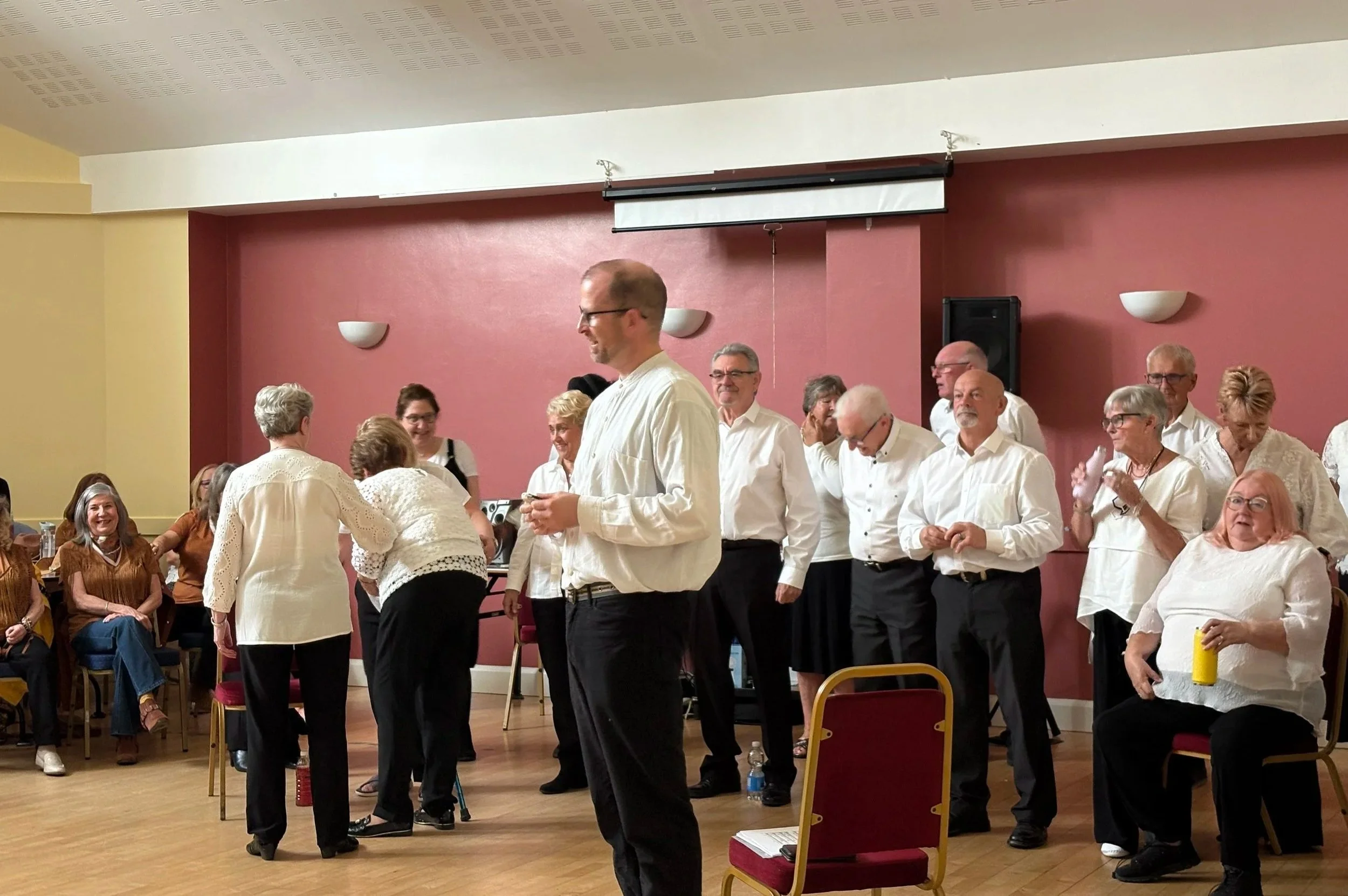 Group of adults gathering in a room with red and beige walls, some standing and some sitting, engaged in conversation or enjoying an event.