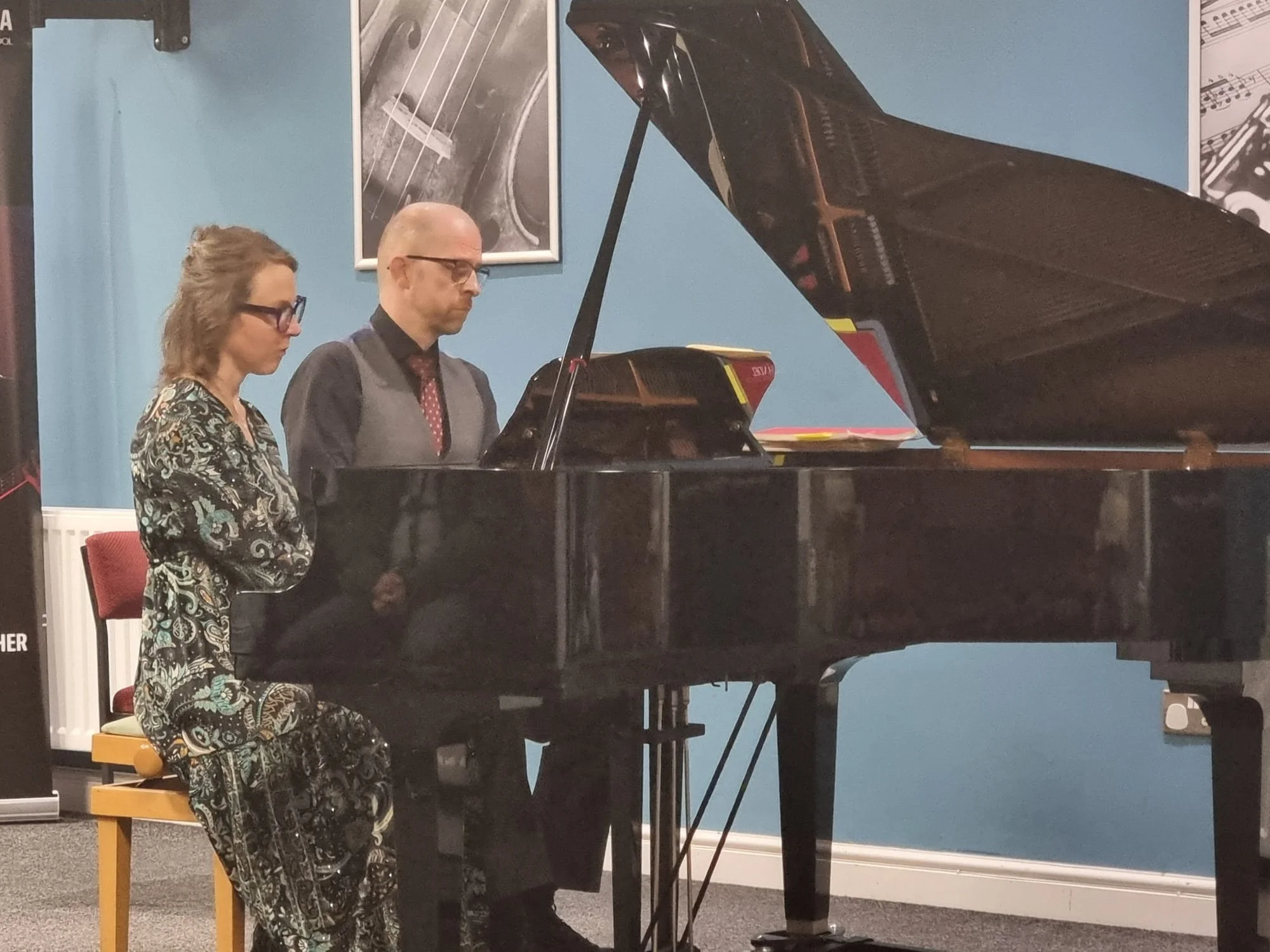 A man and a woman playing the grand piano together. The woman is sitting on a yellow bench, and the man is standing beside her. They are in a room with blue walls and framed pictures, and the scene appears to be a musical performance or practice.
