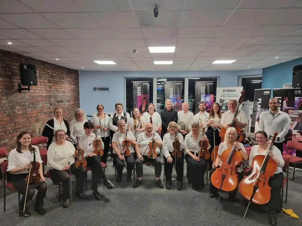 Orchestra of musicians with string instruments, including violins, a cello, and a double bass, posing in a room with brick and blue walls, and banners for Yamaha music products.