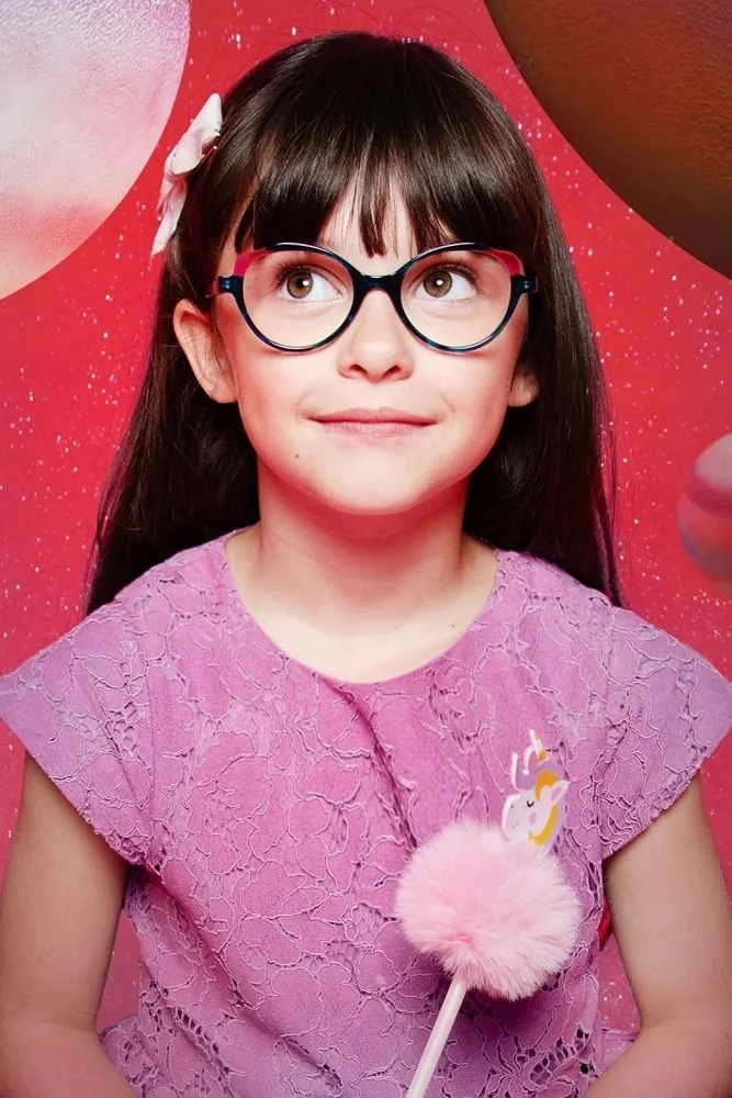 Jeune fille portant des lunettes rondes, une robe rose en dentelle, une barrette blanche dans les cheveux, et tenant une sucette en forme de licorne. Fond rose avec des motifs.