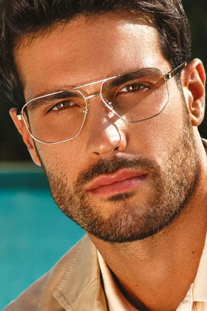 Premier plan d'un homme avec lunette de soleil à l'assemblage métallique, barbe bien taillée, et expression sérieuse, en extérieur.