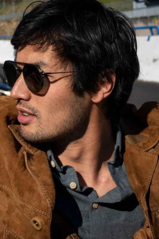 Un jeune homme avec des cheveux foncés et une barbe, portant des lunettes de soleil, une chemise grise et une veste en cuir marron.