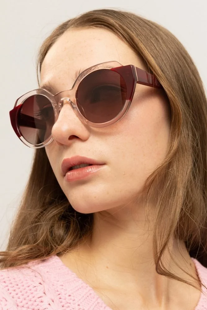Femme avec des lunettes de soleil roses, portant un pull rose, cheveux longs et bruns, fond neutre.