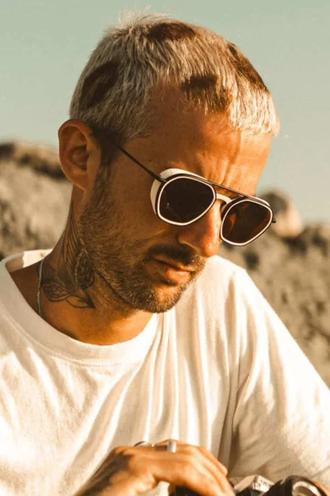 Un homme portant des lunettes de soleil, un t-shirt blanc, avec des tatouages visibles sur le cou, en extérieur dans un environnement ensoleillé.