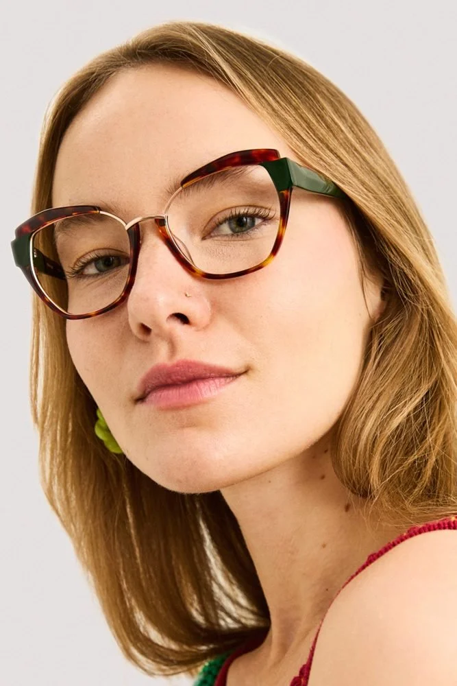 Une jeune femme portant des lunettes à monture tachetée, avec des cheveux châtains clairs, légèrement ondulés, et une peau claire. Elle regarde vers l'appareil avec une expression neutre et porte un haut rouge côtelé.