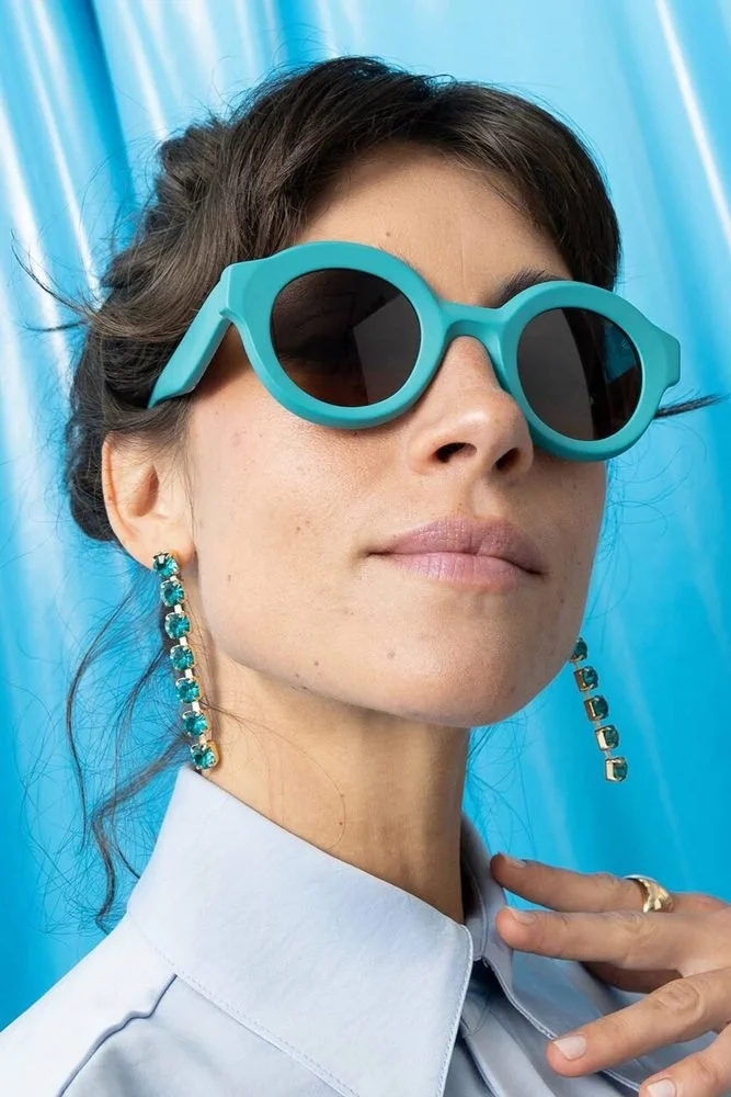 Femme portant des lunettes de soleil bleues, de longues boucles d'oreilles avec des pierres turquoise, et une chemise blanche, avec un fond bleu brillant.