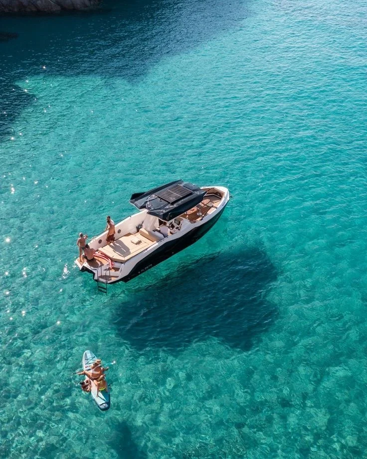 Bateau motorisé avec plusieurs personnes à bord, naviguant dans une eau turquoise claire, avec une personne en paddle devant le bateau.